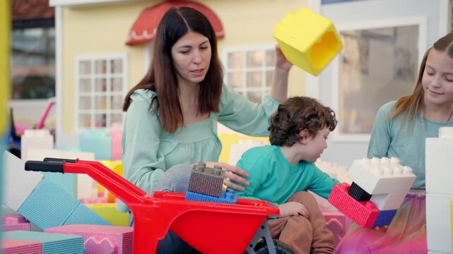 Smiling young woman and two kids playing in game center while putting colorful foam plastic blocks in the wheelbarrow. Slow motion