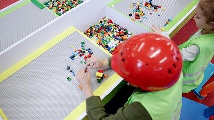 Boy in safe helmet and girl wearing waistcoats playing with colorful toy blocks in game center. Top view. Slow motion. Childhood, entertainment, profession concept © Vlad