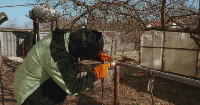 Skilled worker in a protective welding mask and gloves using an electrode to connect metal pipes for a fence in a backyard, creating bright sparks and smoke during the construction process.
