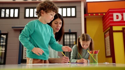 Happy cute little boy sitting at the table and drawing with her mother and sister at game center. Slow motion. Childhood, art, family concept © Vlad
