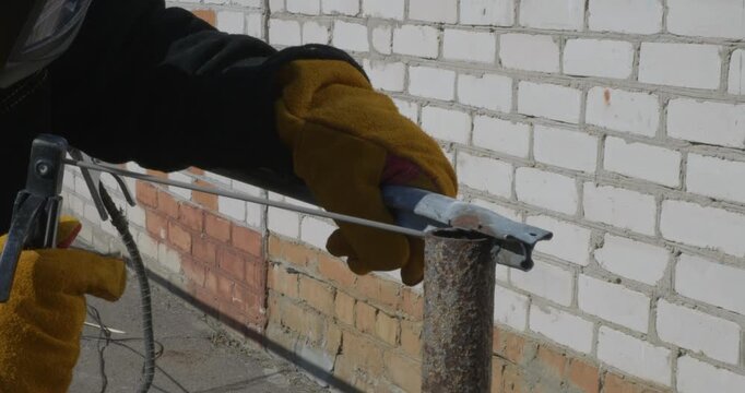 Close up view of a worker in protective gloves welding a metal pipe to a fence post at a construction site, creating bright sparks and smoke against a brick wall background.