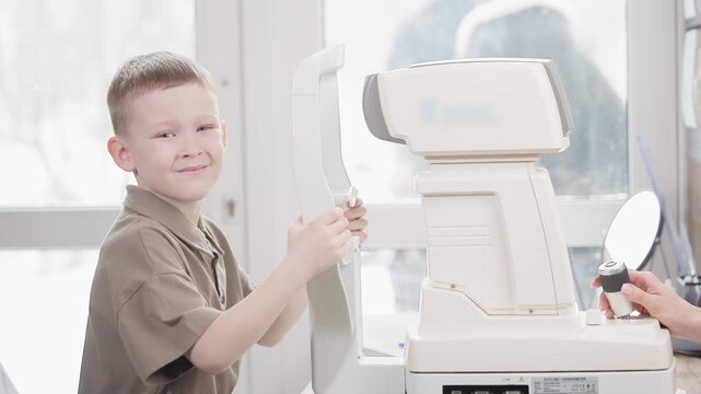 Tracking shot of young boy smiling during eye exam with autorefractor in bright optometry clinic by window.