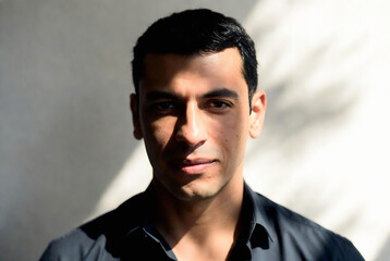 Obraz premium A man with dark hair and a serious expression is looking directly at the camera. He’s standing in front of a plain white wall with strong shadows, suggesting an indoor setting.