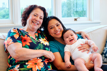 Portrait of senior Caucasian woman, Asian young mother, baby smiling while sitting together on sofa, showing multigenerational family bonding. Ideal for parenting, family care, support campaigns