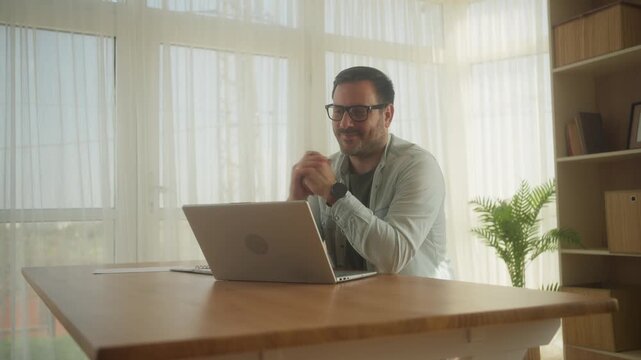 Handsome Caucasian Man Having a Video Call on Laptop Computer while Sitting Behind Desk in Living Room. Freelancer Working From Home and Talking to Colleagues and Clients Over the Internet.