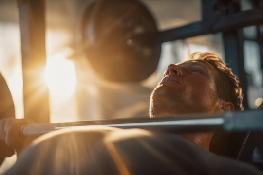 Focused man lifting weights on a bench press in a gym with sunlight streaming through windows during early morning workout session