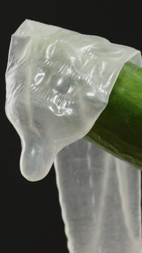 Vertical video. Close-up of a green cucumber partially covered with a transparent latex condom against a black background, showcasing texture and detail with dramatic lighting Safe Sex Education