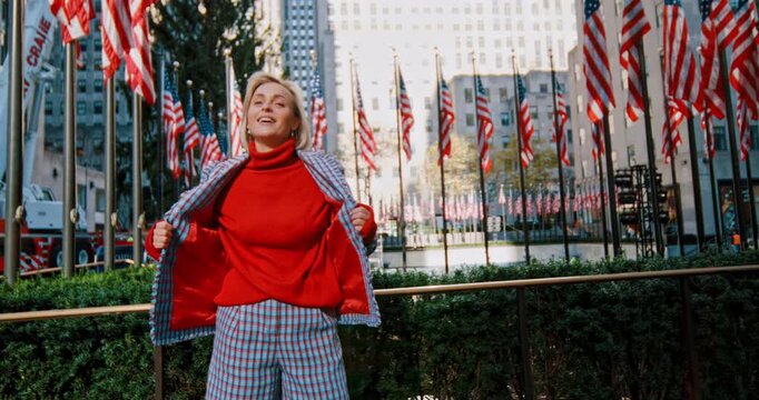 Smiling blonde woman in checkered blue suit. Lady wraps herself in her jacket and looks back at the multiple American flags.
