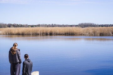 Fototapeta premium Back view of family with father and daughters standing by lake and looking at water together. Warm moment of connection. Concept of family bonding, parenting and peaceful outdoor lifestyle in Poland.