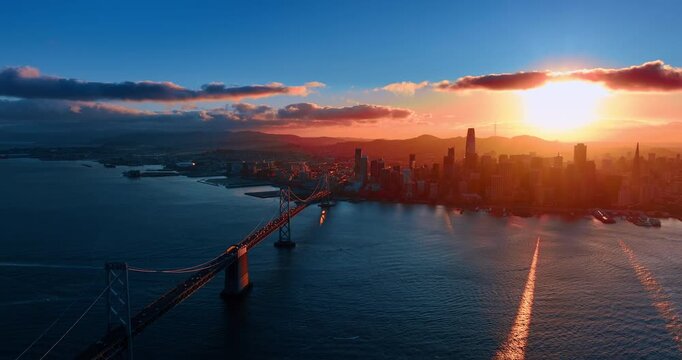 Multiple cars move by the Oakland Bay Bridge to the city center. San Francisco skyline is dazzled by the setting sun.