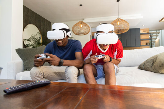 Playing Diverse male friends using VR headsets on sofa in living room, with gaming controllers