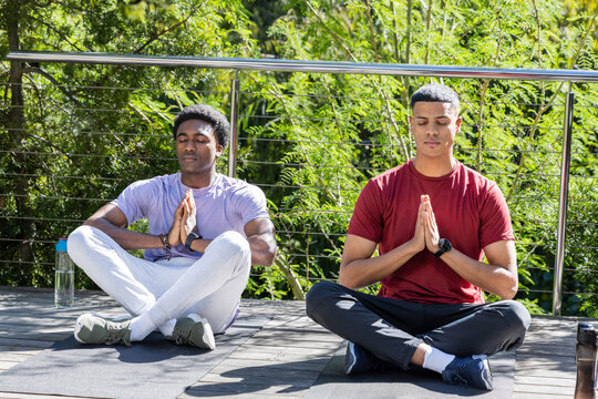 Meditating diverse male friends sitting cross-legged on terrace, with yoga mats and water bottles