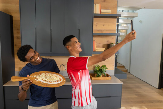 Smiling diverse friends standing in modern kitchen, holding wooden pizza peel and smartphone