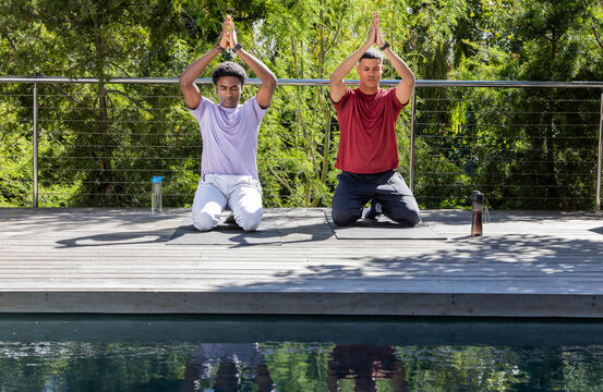 Diverse male friends kneeling in prayer on outdoor deck by pool, with yoga mats, water bottles