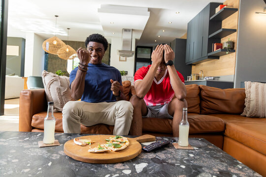 Reacting diverse male friends watching game on couch in living room, with pizza board and remote