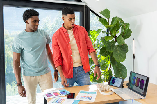Leaning Diverse male coworkers inspecting color swatches on desk in loft, with laptop computer