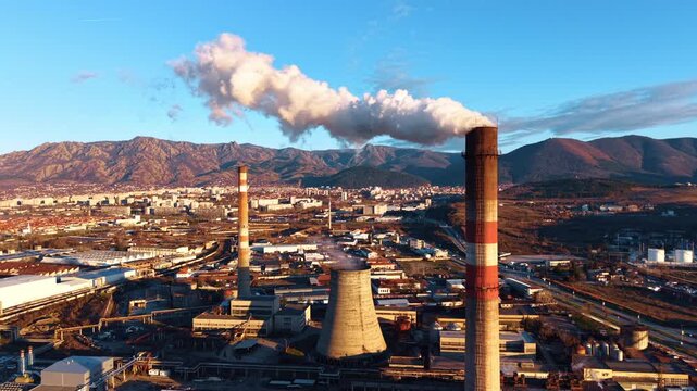 Industrial power plant with smoking chimneys and mountain background. Industrial landscape featuring cooling tower and tall chimneys emitting smoke near city at base of mountains.