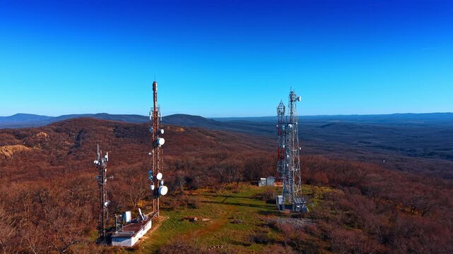 High angle view of communication towers on forested hill. Group of metal lattice towers with satellite dishes and antennas surrounded by trees under blue sky.