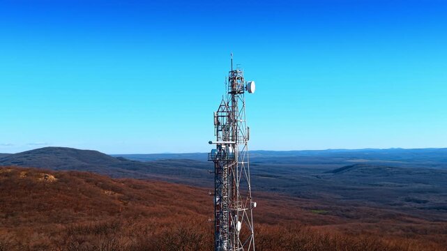 Telecommunication tower on wooded hill under clear blue sky. Metal lattice mast with antennas and satellite dishes on forest ridge with rolling hills.