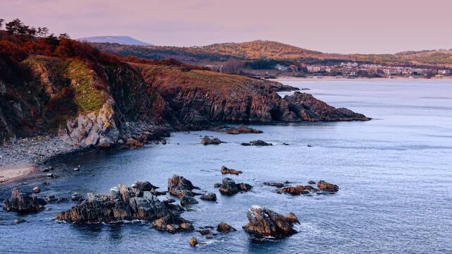 Coastal landscape with rocky shore and distant seaside town. View of jagged rocks in the water along a cliffside with buildings in the background during sunset.