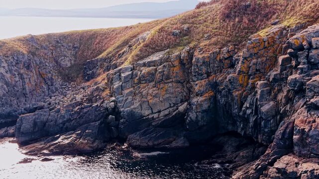 Close up view of coastal rock formations and cliffs. Detailed view of layered grey and brown stone cliffs with sparse vegetation by the sea.