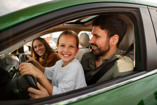 Father teaching little child girl to drive car, daughter holding steering wheel and smiling at camera, family enjoy time together at weekend and driving lesson