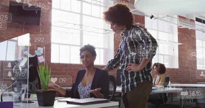 Seated woman pointing at monitor, standing colleague leaning in, icons floating, showing metrics