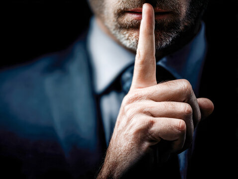 Man in formal suit with finger on lips signaling silence and secrecy in a dimly lit environment focused on privacy and quietness concept