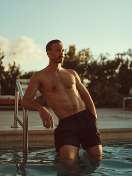 Man in swim trunks leaning on pool ladder at sunset with muscular physique
