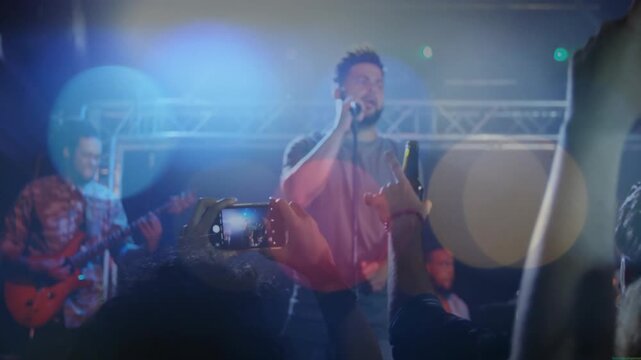 Lead singer singing into mic center stage, audience raising phones recording concert, lens flare
