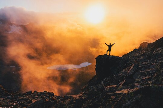 Successful hiker silhouette on mountain peak at sunset, triumphant man standing on rocky cliff above clouds, golden hour mountain landscape