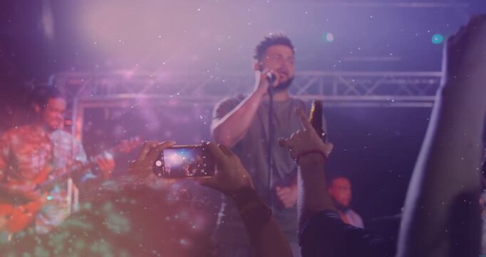 Male vocalist singing concert as fan recording, crowd raising phones, particle overlay over stage
