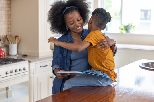 African American mother and child sitting on wooden countertop reading picture book in kitchen