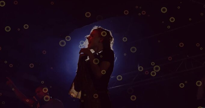 Lead female singer thrusting on beat singing into mic backlit slowing orbs drifting band in dark