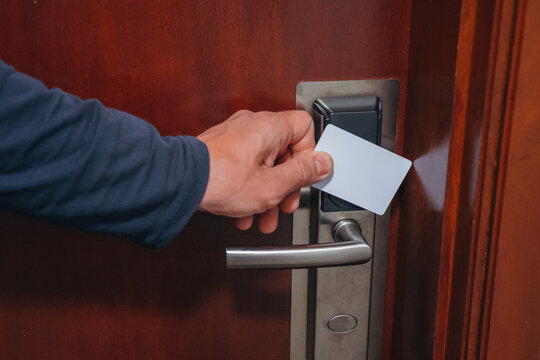 A close-up shot of a person's hand using a white plastic electronic key card to unlock a dark wooden door with a modern silver metal handle and reader.