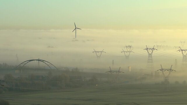 Ethereal aerial perspective of a single wind turbine and high voltage power lines emerging from dense fog during a tranquil, warm sunrise