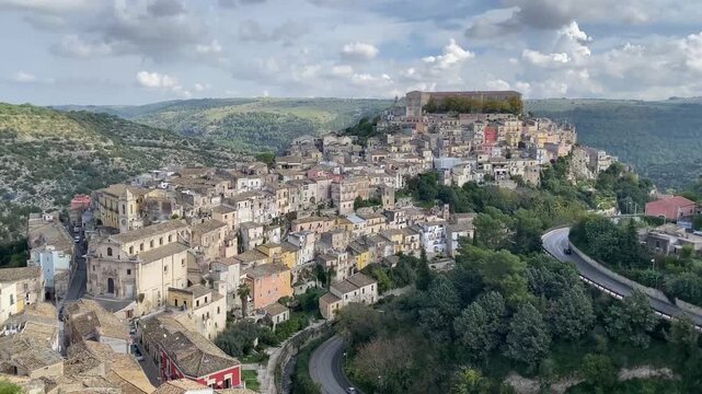 Fixed panoramic view of Modica, Sicily's UNESCO baroque town perched on hills. Colorful clustered buildings, winding roads, green valleys under cloudy skies. Timeless Sicilian landscape