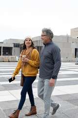 Middle-aged couple walking in plaza, woman holding coffee and smartphone, wearing beanie © wavebreak3