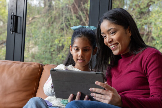 Mother and Indian child sharing tablet on leather sofa beside black-framed window bow visible