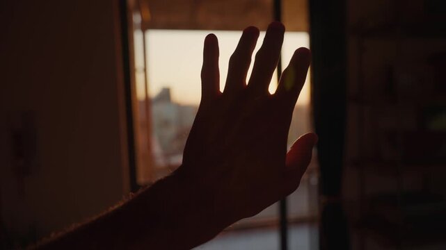 Hand silhouette in front of setting or raising sun in dark room with open door. Sun shining through person's fingers creating lens flare. Hope and green energy. Early morning warm light rays indoors
