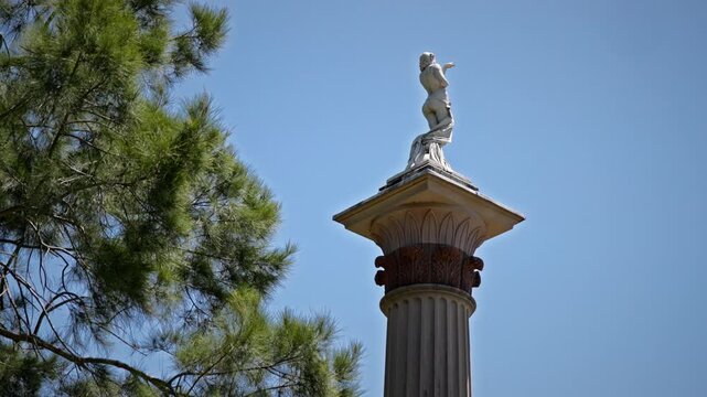 Classical statue atop ornate column in Centennial Park Sydney, framed by pine branches against clear blue sky, evoking heritage, art, and tranquility.