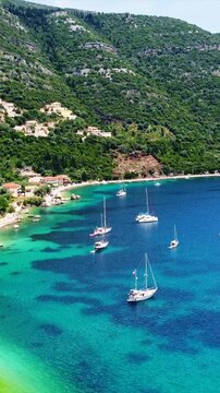 Vertical aerial of a coastal village in Rouda Bay, sailboats anchored in turquoise Ionian waters along a rocky shore with villas on lush green hills, Lefkada, Greece