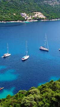 Vertical aerial of sailboats in calm turquoise Rouda Bay at Mikros Gialos Beach, surrounded by lush green mountains, Mediterranean summer scene