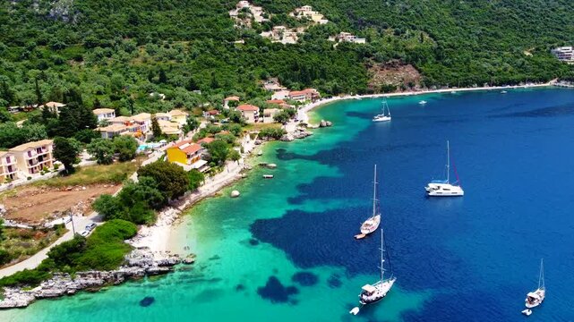 Aerial of coastal village in Lefkada, sailboats anchored in turquoise Ionian waters along a rocky shoreline with lush green hills, Rouda Bay, Greece.