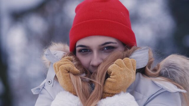 Close up portrait of a young woman wearing yellow gloves and a red beanie, pulling her jacket collar up in a snowy forest.