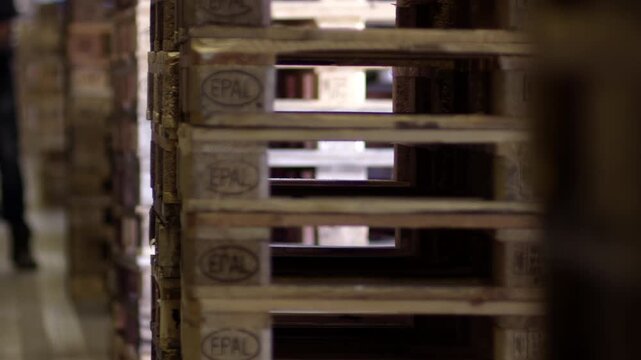 Stacked wooden pallets in a warehouse with a forklift and a worker moving boxes in the background.