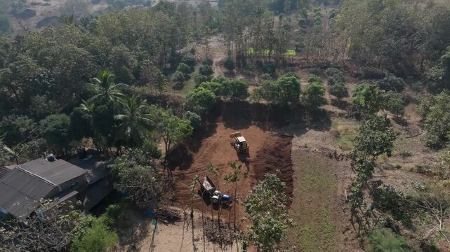 Overhead aerial view of an excavator working in red soil farmland clearing surrounded by tropical trees and small rural buildings in Vansda, Gujarat. Bright daylight with dry season landscape.