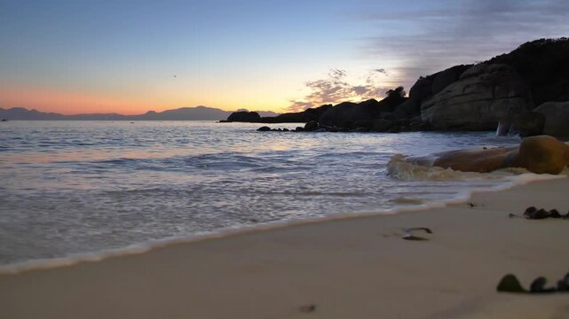 Golden hour light reflects on the wet sand as calm ocean waves roll onto a secluded beach with rocky outcrops and silhouetted mountains in the distance at Millers Point, Simon's Town.