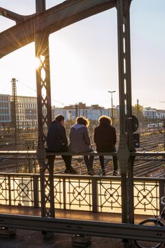 Three youths sitting on the balustrade of the Hackerbr&uuml;cke bridge over the railway tracks looking into the distance, back light, Munich, Upper Bavaria, Bavaria, Germany
