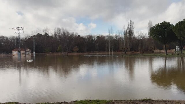 Slow motion shot of Adaja river flooding a park in Avila, showing submerged trees, water reaching huts and electrical lines in an extreme weather situation.